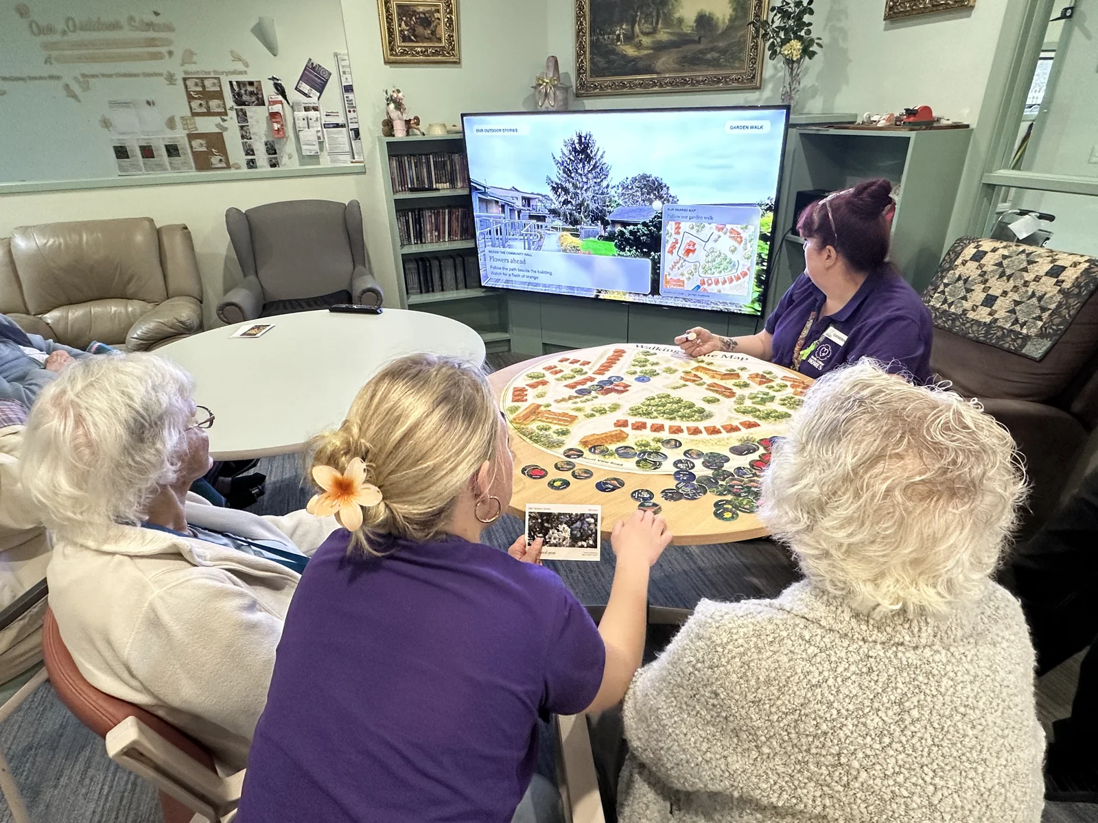 Residents and care staff gathered around a round walking route map beside a screen showing the garden